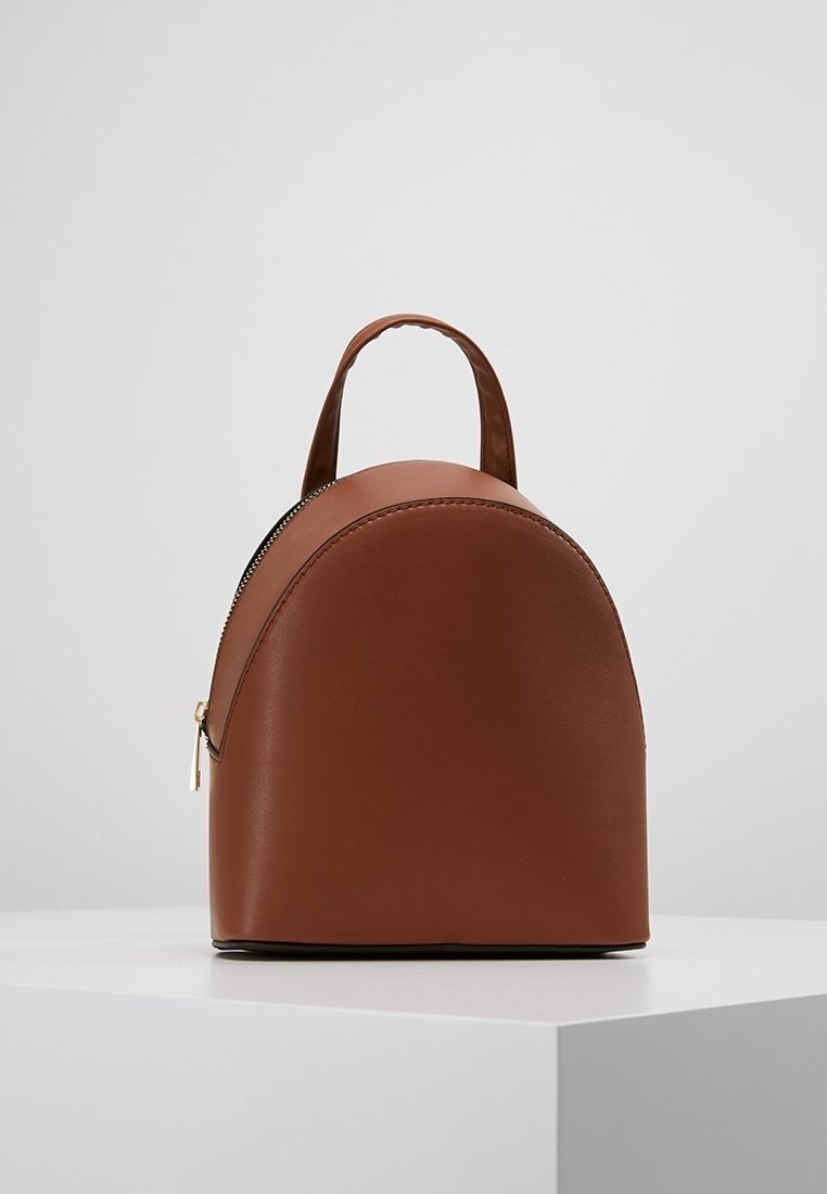 Mochila de cuero marrón con forma redondeada. Cuenta con cierre de cremallera, asa pequeña y textura lisa. Diseño minimalista sin patrones.