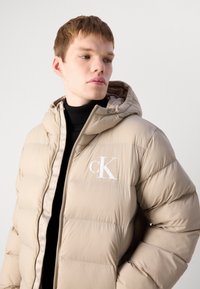 Young man wearing beige puffer jacket with hood and white "cK" logo, looking right, against plain light background.