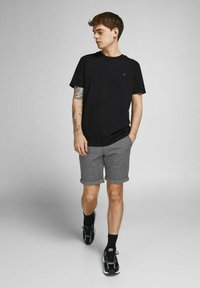 T-shirt noire avec un petit accent rouge, accompagnée de shorts gris avec un ourlet retroussé. Le mannequin porte des baskets noires et des chaussettes mi-chèville.