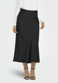 Jupe midi noire avec un ourlet évasé, confectionnée en tissu lisse. Portée avec une blouse blanche et associée à des escarpins à bride cheville gris.