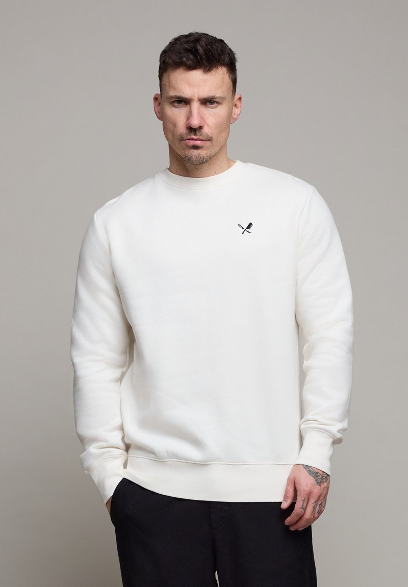 Homme aux cheveux courts portant un sweat-shirt blanc avec un petit logo de batte et de bâton croisés, debout devant un fond gris uni.