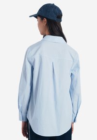 Camicia a righe azzurro chiaro con colletto a punta, maniche lunghe e polsini abbottonati, con yoke e dettagli a piega sul retro.