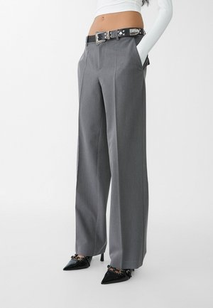 Femme portant un pantalon gris taille haute à jambes larges avec ceinture noire et chaussures noires à talons aiguilles pointues, haut blanc à manches longues coupé au-dessus de la taille.