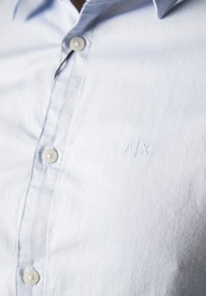 Camicia azzurra chiaro, con bottoni e tessuto morbido e strutturato. Presenta un piccolo logo ricamato "A/X" e bottoni rotondi cuciti.
