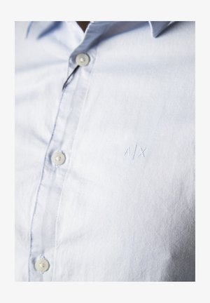 Camicia azzurra chiaro, con bottoni e tessuto morbido e strutturato. Presenta un piccolo logo ricamato "A/X" e bottoni rotondi cuciti.