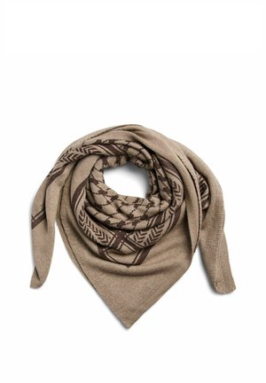 Jeune personne aux cheveux tressés portant un pull vert et une écharpe beige à motifs nouée autour du cou sur un fond uni.