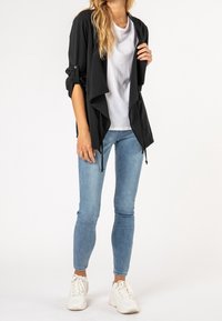 Femme portant une veste noire ouverte à l'avant, un t-shirt blanc, un jean skinny bleu clair et des baskets blanches, debout devant un fond blanc.