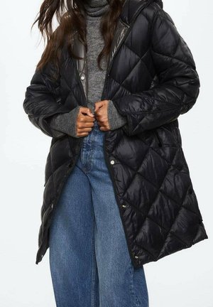 Femme boutonnant un long manteau noir matelassé par-dessus un pull à col roulé gris et un jean bleu à jambes larges.
