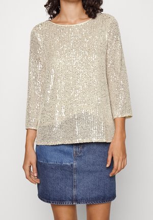 Femme portant un haut beige à sequins à manches longues associé à une mini-jupe en denim bleu bicolore, visible du cou jusqu'à mi-cuisse.