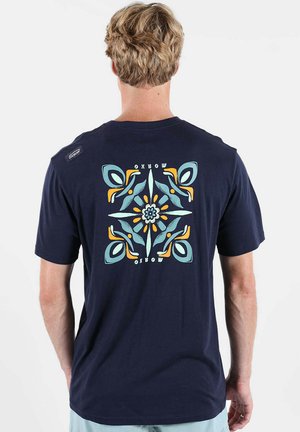 Homme portant un t-shirt bleu marine avec un motif floral et de feuilles symétrique en bleu, bleu sarcelle et orange dans le dos, portant la mention "OXBOW".