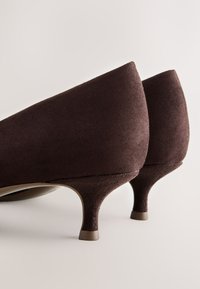 Paire de chaussures en daim marron foncé à talons kitten, vue de dos sur un fond clair et neutre.