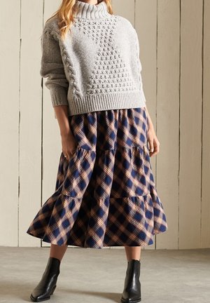 Grå stickad tröja med hög krage. Mörkblå rutigt kjol med tan accenter, med en lagerdesign. Svarta ankelstövlar fulländar looken.