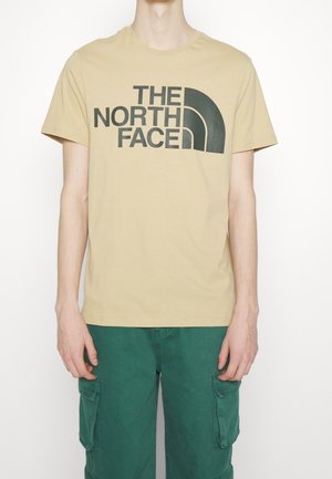 Beżowa koszulka z bawełny z dużym czarnym logo „THE NORTH FACE” z przodu. Krótkie rękawy i klasyczny okrągły dekolt.