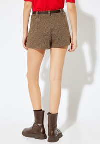 Pantalones cortos de patrón espiga marrón con cintura alta, combinados con botas de tobillo brillantes marrones y una camisa roja.