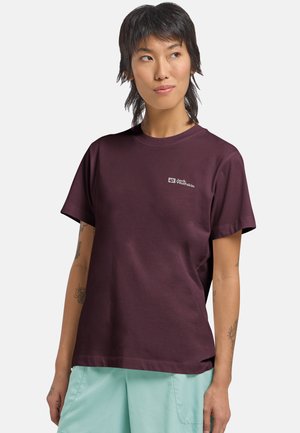 Persona con capelli corti e scuri che indossa una maglietta maroon di Jack Wolfskin e pantaloni azzurri chiari, in piedi su uno sfondo bianco semplice.