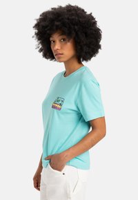 Turquoise cotton t-shirt with a crew neck, featuring a small, colorful palm tree graphic on the chest. Short sleeves, relaxed fit.
