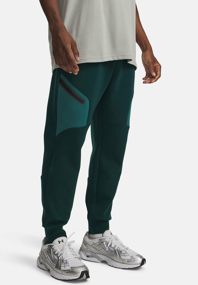 Dunkelgrüne Jogginghosen mit Akzentpaneelen, seitlichen Reißverschlusstaschen und schmaler Passform. Kombiniert mit silbernen Sportschuhen.