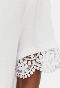 T-shirt bianco con maniche a lunghezza gomito, caratterizzato da un intricato bordo in pizzo bianco. Il pizzo presenta motivi floreali e piccoli dettagli rotondi.