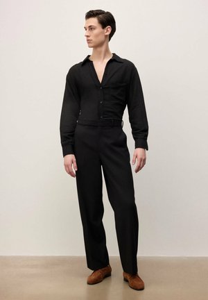 Jeune homme debout, portant une chemise noire à boutons, un pantalon noir taille haute et des chaussures en daim marron, regardant vers sa gauche sur un fond uni.