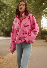 Fleece-Pullover in leuchtendem Pink mit Kirsch- und Schleifenmuster, ausgestattet mit einem Reißverschlusskragen und lockerer Passform. Kaum sichtbare Hintergrundlandschaft.