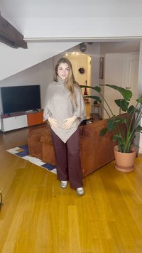 Jovem mulher de pé na sala de estar a usar um poncho bege, calças bordô e sapatos prateados, com um sofá castanho e uma grande planta verde nas proximidades.