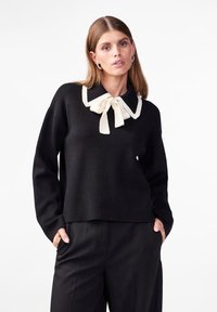 Pull noir avec un large col blanc agrémenté d'un nœud. Tissu doux, coupe ample et manches longues. Associé à un pantalon noir.