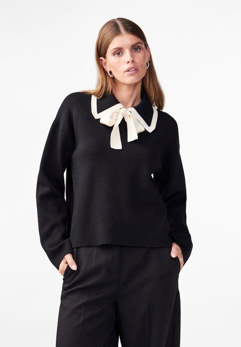 Pull noir avec un large col blanc agrémenté d'un nœud. Tissu doux, coupe ample et manches longues. Associé à un pantalon noir.