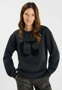 Svart sweatshirt i mjukt material, med en stor, matt logotyp på framsidan. Kombineras med mönstrade, mörka leopardmönstrade byxor.