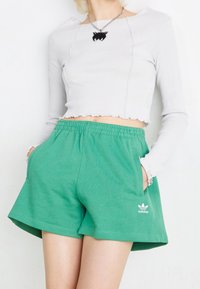 Žena v zelených šortkách Adidas s kapsami a bílém dlouhém cropped topu, stříbrném řetízku s černým přívěskem.