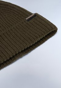 Stickad olivgrön beanie med ribbad textur och en liten svart etikett på sidan, med varumärkesnamnet i kontrasterande färger.