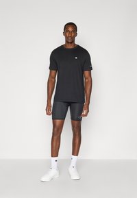 Zwarte katoenen t-shirt en grijze shorts met logo-details. Model draagt witte sneakers en sokken, staand tegen een effen achtergrond.