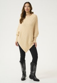 Beige gebreide poncho met een gestructureerd zigzagpatroon, V-hals en oversized mouwen, gecombineerd met zwarte slim fit jeans en zwarte cowboylaarzen.