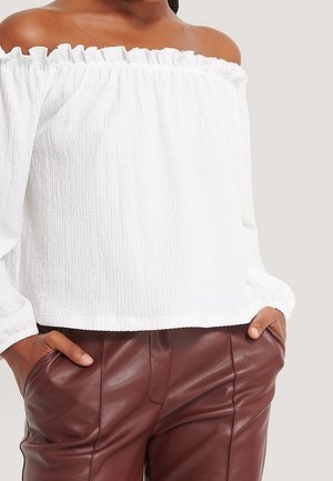 Hvid off-the-shoulder top med struktureret stof, flæsekrave og lange ærmer, parret med brune læderbukser.