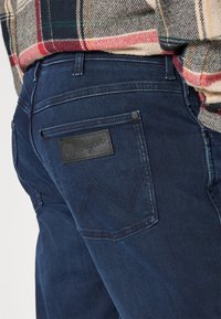 Jeans de denim navy cu un patch din piele cu logo-ul Wrangler pe buzunarul din spate. Prezintă cusături de contrast și un design tipic cu cinci buzunare.