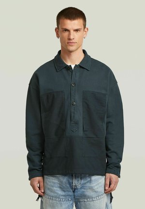 Giovane uomo con capelli corti che indossa un pullover verde scuro a maniche lunghe con bottoni e jeans azzurri su uno sfondo semplice.
