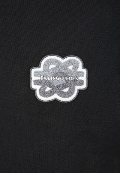 Šedé a bílé texturované vyšívané logo s propletenými tvary a textem "FILLING PIECES" na černé látce.