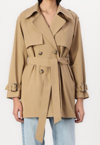 Khaki trenchcoat met dubbele rij knopen, brede kraag, ceintuur aan de taille, en opgerolde mouwen met gespaccenten. Gladde textuur.