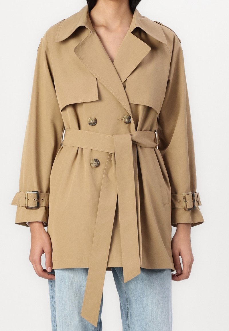 Khaki trenchcoat met dubbele rij knopen, brede kraag, ceintuur aan de taille, en opgerolde mouwen met gespaccenten. Gladde textuur.