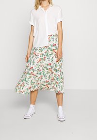 Chemise blanche à manches courtes avec des boutons, associée à une jupe fluide à imprimé floral crème, ornée de fleurs colorées et de feuillage. Baskets blanches.