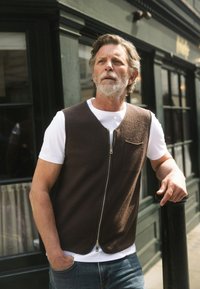 Smeđi prsluk od flisa s prednjim patentnim zatvaračem, nošen preko bijele majice kratkih rukava. Ima džep na prsima i zaobljeni rub, postavljen na tamnozelenoj pozadini.