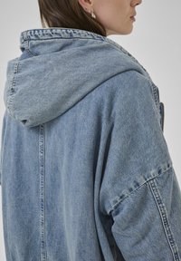 Giubbotto di jeans in azzurro chiaro con cappuccio morbido, fit oversize, cuciture a taglio vivo e dettagli di cucitura visibili.