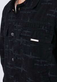 Chaqueta negra con un patrón sutil de estrellas y texto "PXP" en todo el diseño. La tela tiene una textura suave, con bolsillos delanteros y cierres de botones a presión.