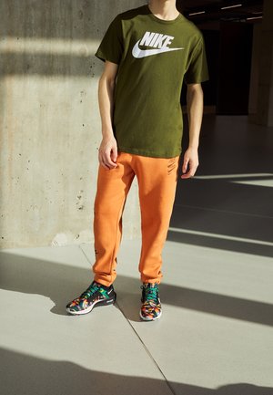 Homme portant un t-shirt Nike vert olive, un pantalon de survêtement Nike orange et des baskets Nike colorées, debout à l'intérieur sur un sol clair.