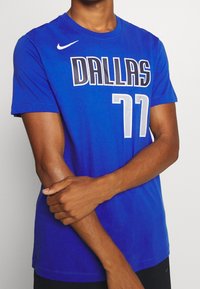Persona che indossa una maglia da basket blu dei Dallas con il numero 77 e un logo Nike bianco, con le braccia incrociate, in piedi contro uno sfondo semplice.