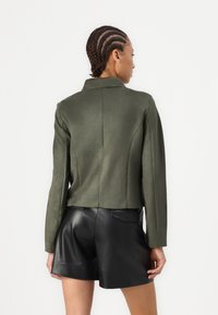 Olivengrønn cropped jakke med strukturerte skuldre, glatt tekstur og en enkelt krage. Kombinert med svarte skinnshorts.