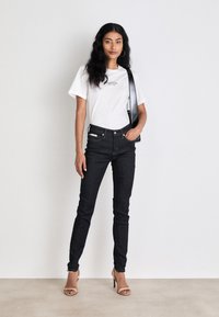 Temno modri skinny jeans z visokim pasom, v kombinaciji z belo bombažno majico z logotipom, dopolnjeni z natikači v nude barvi.