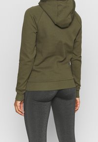 Olijfgroen hoodie met een capuchon, ribgebreide cuffs en zoom; gecombineerd met donkergrijze leggings, die een gladde, gestructureerde stof laten zien.
