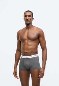 Szürke pamut boxernadrág fehér gumírozott derékpánttal, amelyen a "GANT" márkanév tengerészkék színnel van nyomtatva. A modell semleges testtartásban áll.