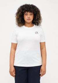 Λευκό βαμβακερό T-shirt με κοντά μανίκια, στρογγυλή λαιμόκοψη και μικρό λογότυπο "CK" σε μπορντό στο αριστερό στήθος. Απλός σχεδιασμός, λεία υφή.