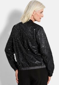 Veste bomber noire avec un revêtement en sequins, des poignets et un ourlet côtelés. Texture lisse avec des détails scintillants et un design de dos arrondi.